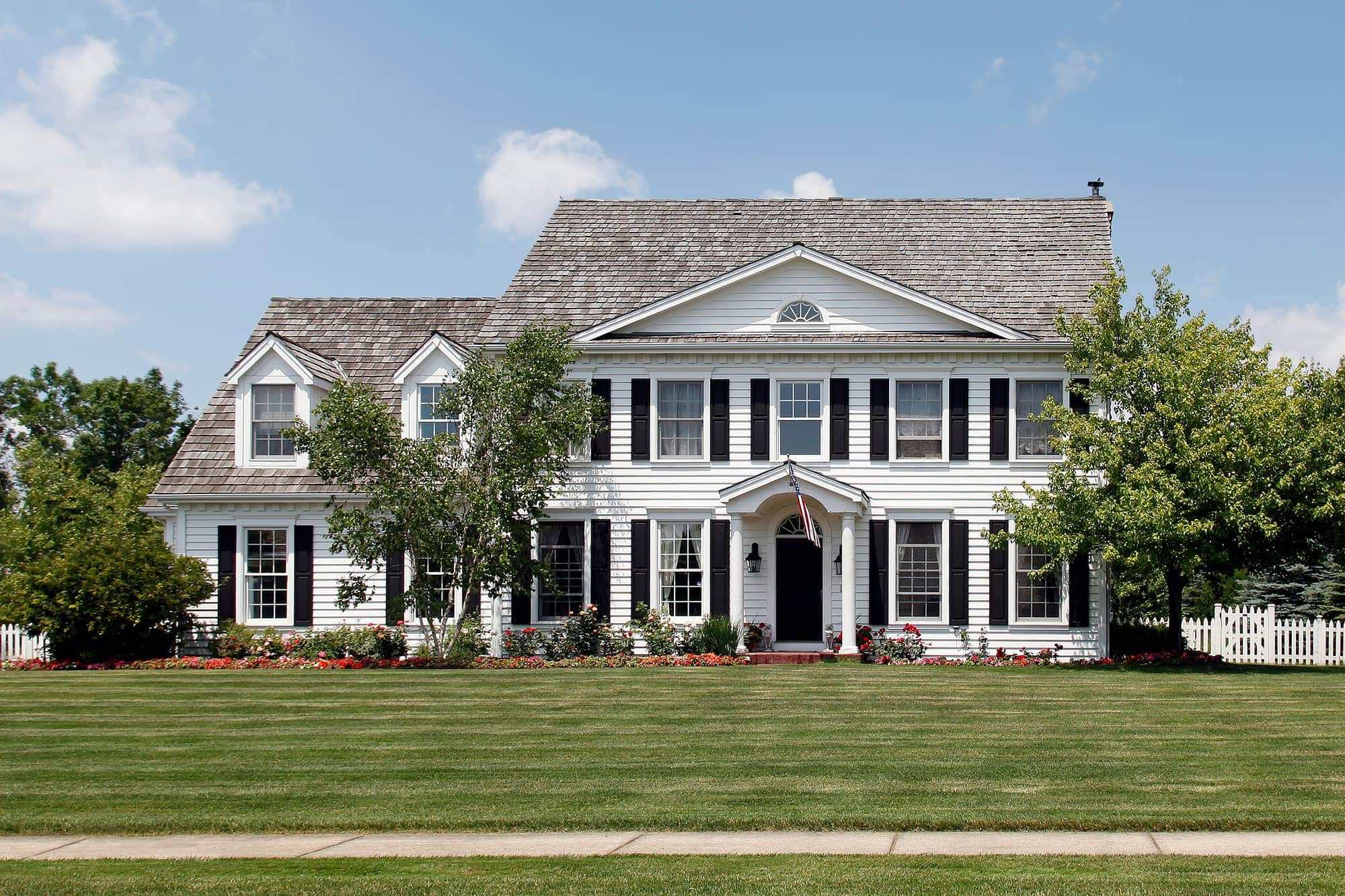 Two-story white colonial house surrounded by trees and flowers